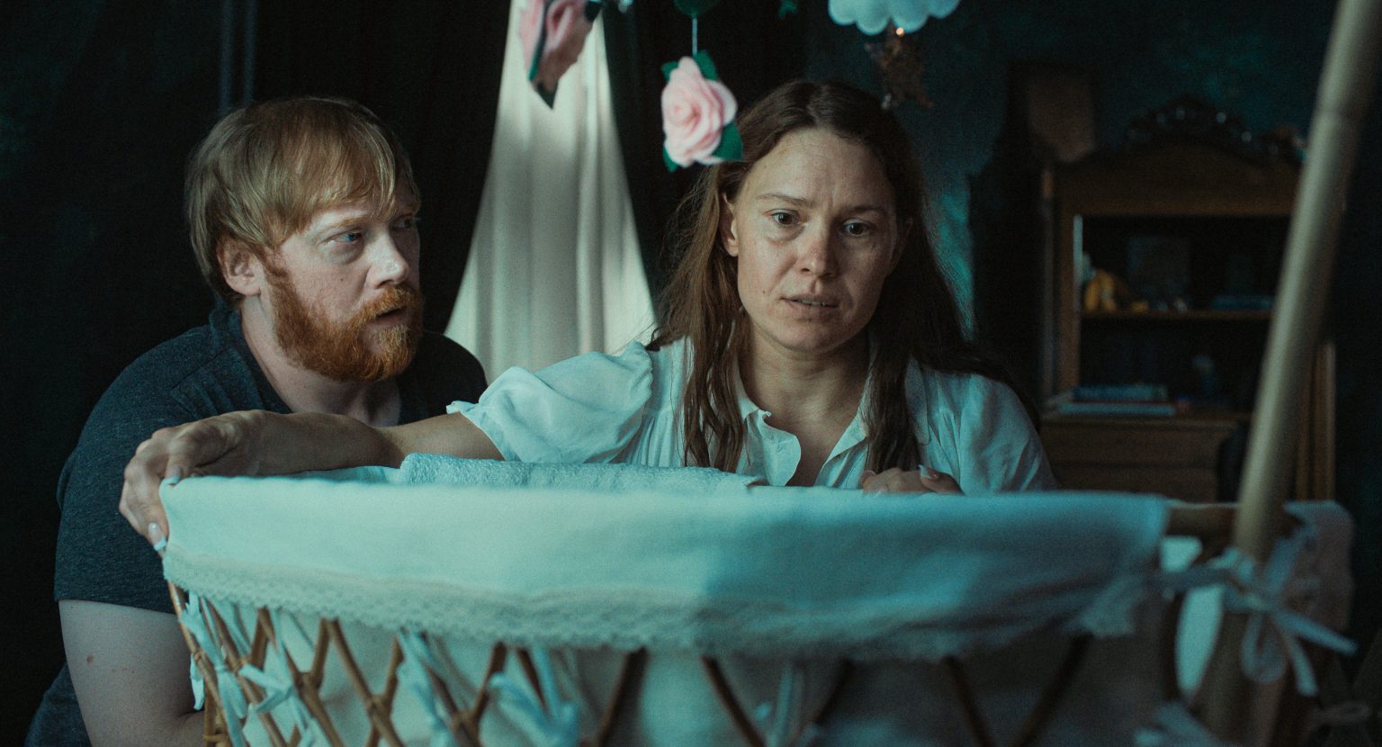 A man and woman look with concern into a white bassinet in a dimly lit room, with flower decorations hanging above.