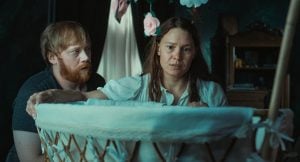 ‘Nightborn’ Review – A Nightmare On Motherhood A man and woman look with concern into a white bassinet in a dimly lit room, with flower decorations hanging above.