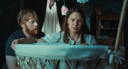 ‘Nightborn’ Review – A Nightmare On Motherhood A man and woman look with concern into a white bassinet in a dimly lit room, with flower decorations hanging above.