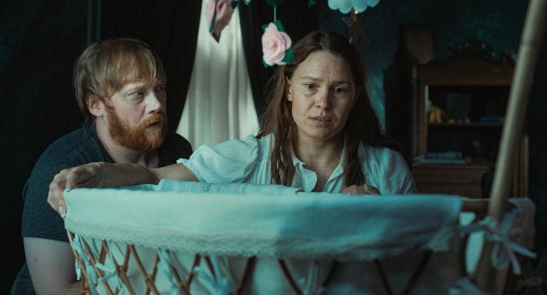‘Nightborn’ Review – A Nightmare On Motherhood A man and woman look with concern into a white bassinet in a dimly lit room, with flower decorations hanging above.