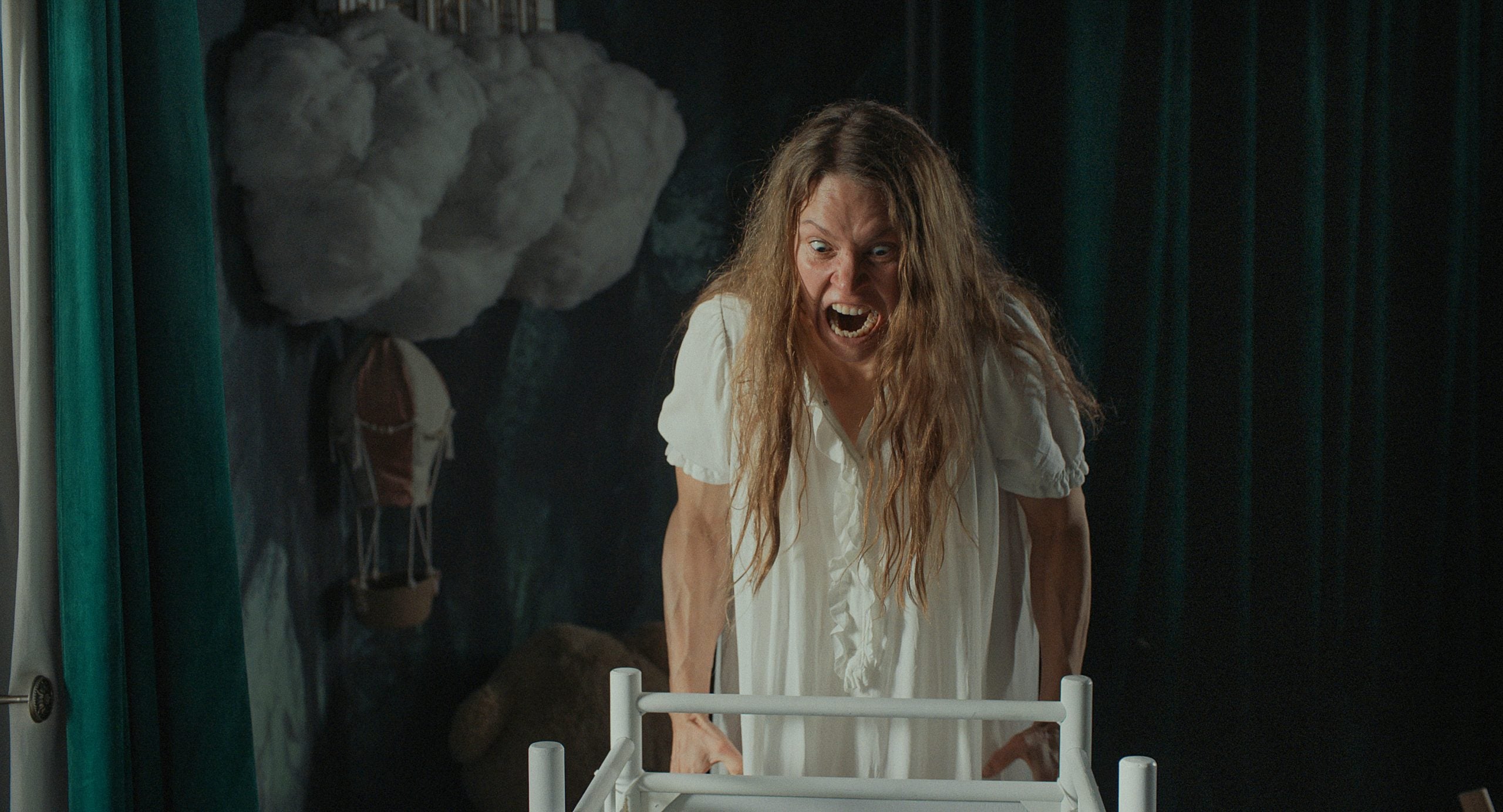 A woman in a white nightgown leans over a white crib, screaming with an angry expression in a dimly lit room decorated with clouds.