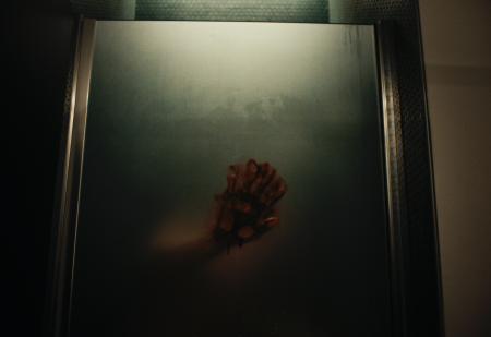 A red handprint and blurred arm are visible on the fogged glass door of a shower, with dim lighting above.