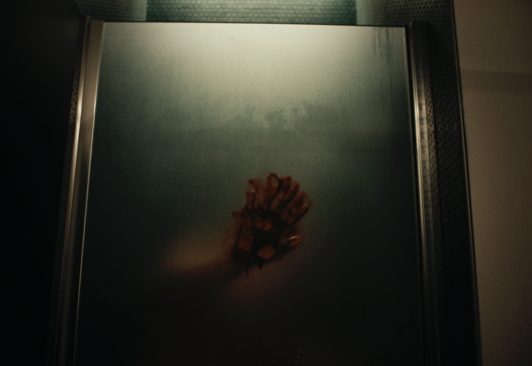 A red handprint and blurred arm are visible on the fogged glass door of a shower, with dim lighting above.