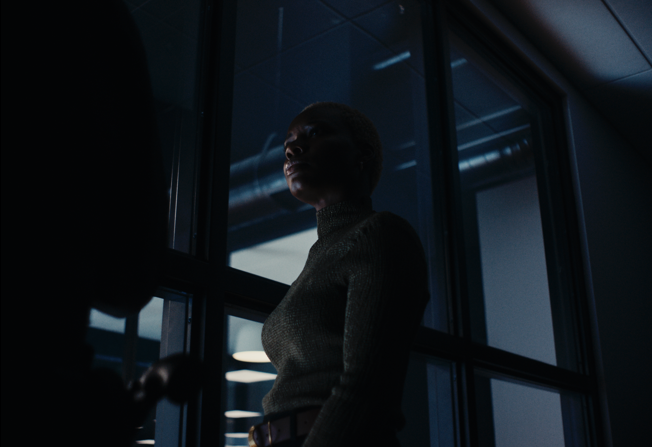 A person stands indoors near large windows at night, illuminated by dim, cool lighting and reflections from outside.