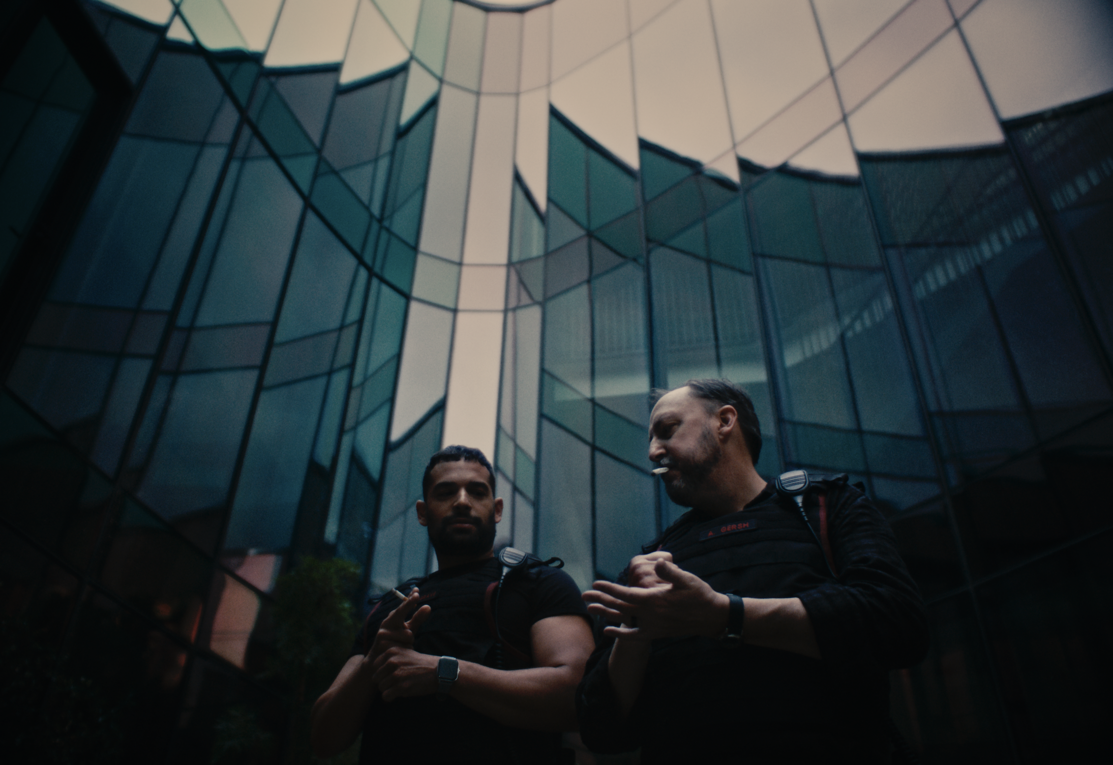Two men in black tactical gear stand outside a modern glass building, engaged in conversation, with reflective windows visible behind them.