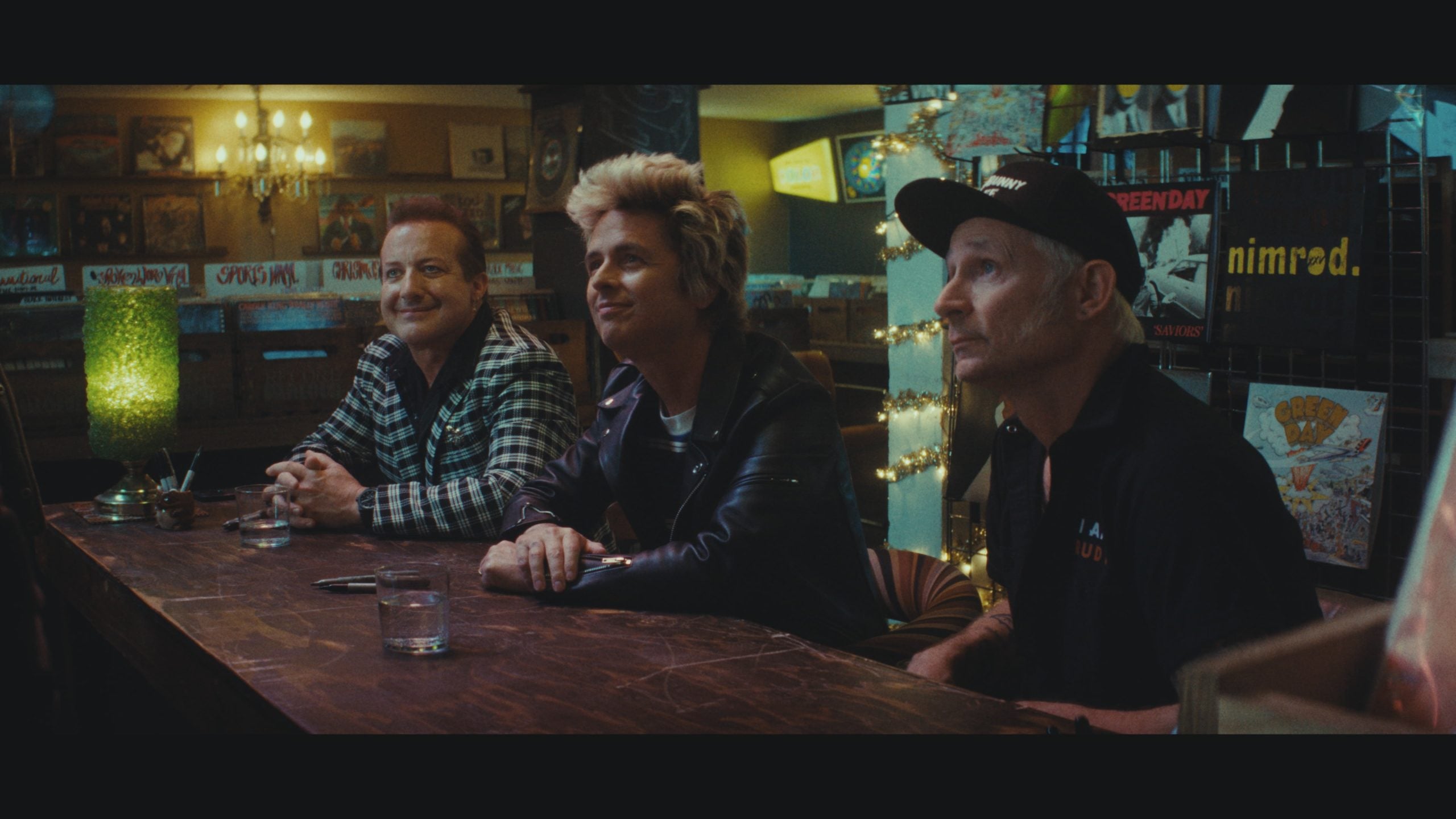 Three men sit at a wooden bar in a dimly lit record store, each with a glass of water in front of them; vinyl records and posters are visible in the background.