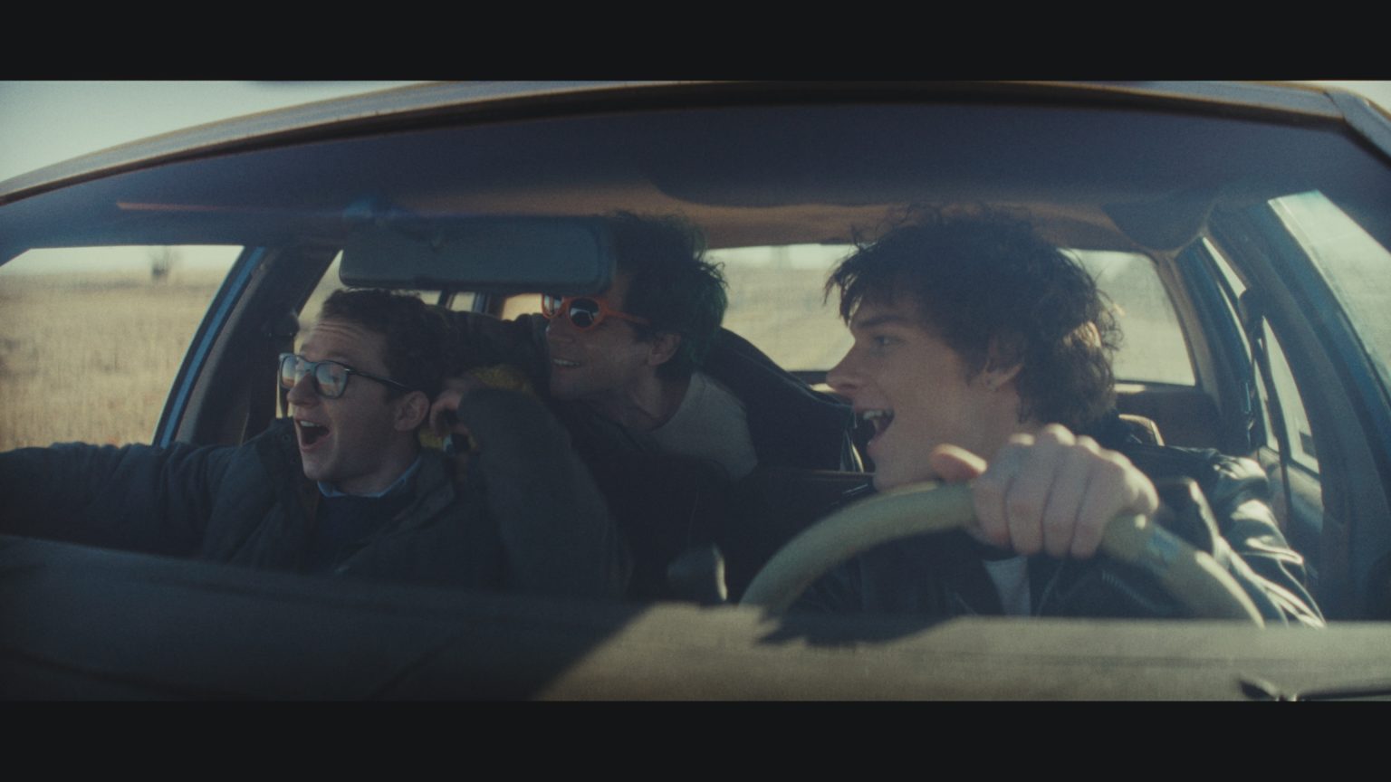 ‘Nimrods’ Review – A Charming if Somewhat Middling Coming-of-Age Comedy Three young men are in a moving car; one drives while the other two ride as passengers. They are smiling or laughing, and sunlight streams in through the windshield.