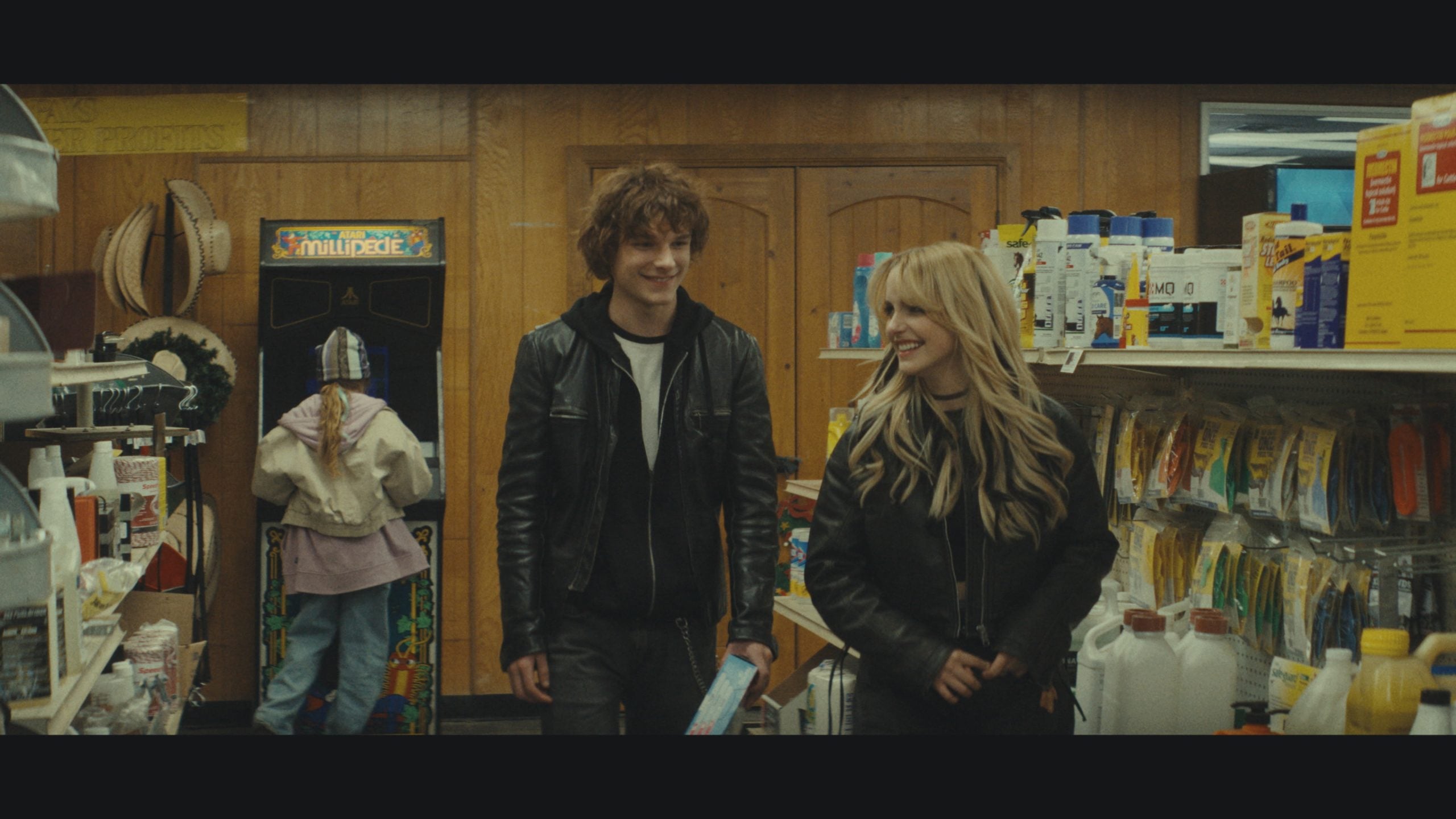 Two young adults walk through a store aisle, smiling and talking, while another person plays at an arcade machine in the background. Shelves are stocked with various household items.