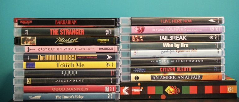 A stack of 16 Blu-ray and DVD movie cases are arranged upright in two columns against a teal background. The visible spines display various titles and cover designs.