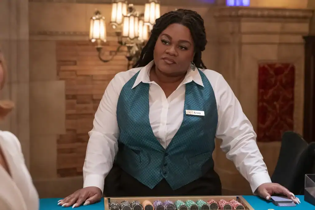 A casino dealer in a teal vest stands behind a gaming table, looking ahead. Stacks of chips are arranged in front of her.