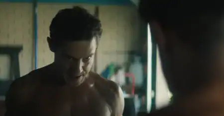 Shirtless man with a tense expression looks at his reflection in a mirror, indoor setting with blurred background items.