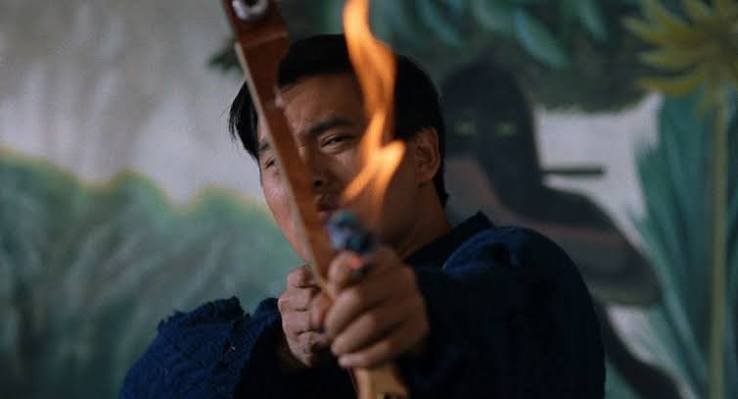 A man aims a bow with a flaming arrow indoors, with a mural featuring a dark figure and greenery in the background.
