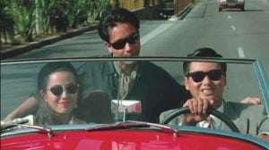 Three people wearing sunglasses ride in a red convertible on a sunny day; two are in front, one is standing behind them, leaning over the seat.