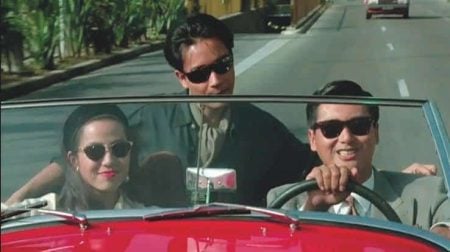 Three people wearing sunglasses ride in a red convertible on a sunny day; two are in front, one is standing behind them, leaning over the seat.