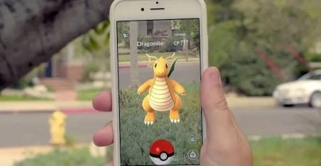 A person holds a phone displaying an augmented reality game with a Dragonite Pokémon appearing on the screen outdoors.