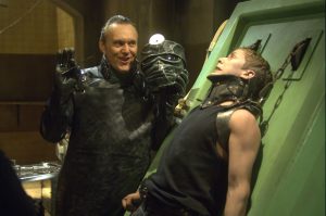 A man in dark protective gear holds a futuristic helmet, while another man is restrained and leaning back against a green metal platform with a distressed expression.