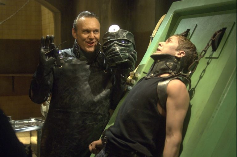 Lionsgate To Release Cult Horror Musical ‘Repo! The Genetic Opera’ On 4K UHD Blu-Ray This October A man in dark protective gear holds a futuristic helmet, while another man is restrained and leaning back against a green metal platform with a distressed expression.