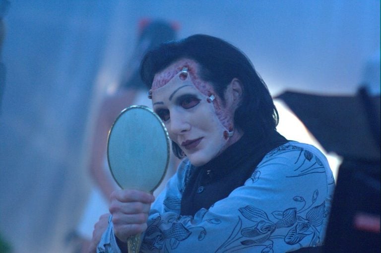 Person with theatrical makeup, showing exposed skin and stitches, holds a hand mirror and looks at their reflection in a dimly lit room.
