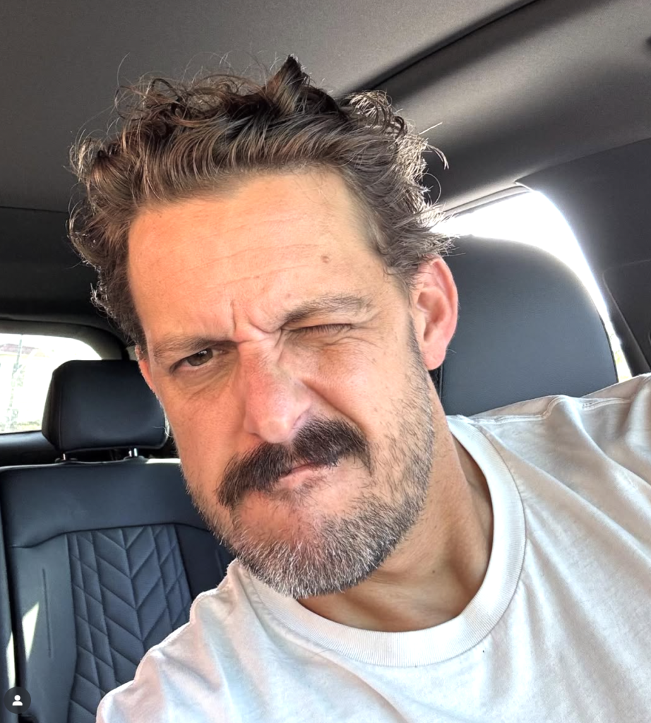 A man with curly hair and a mustache sits in a car, wearing a white T-shirt and squinting one eye while making a slight grimace.