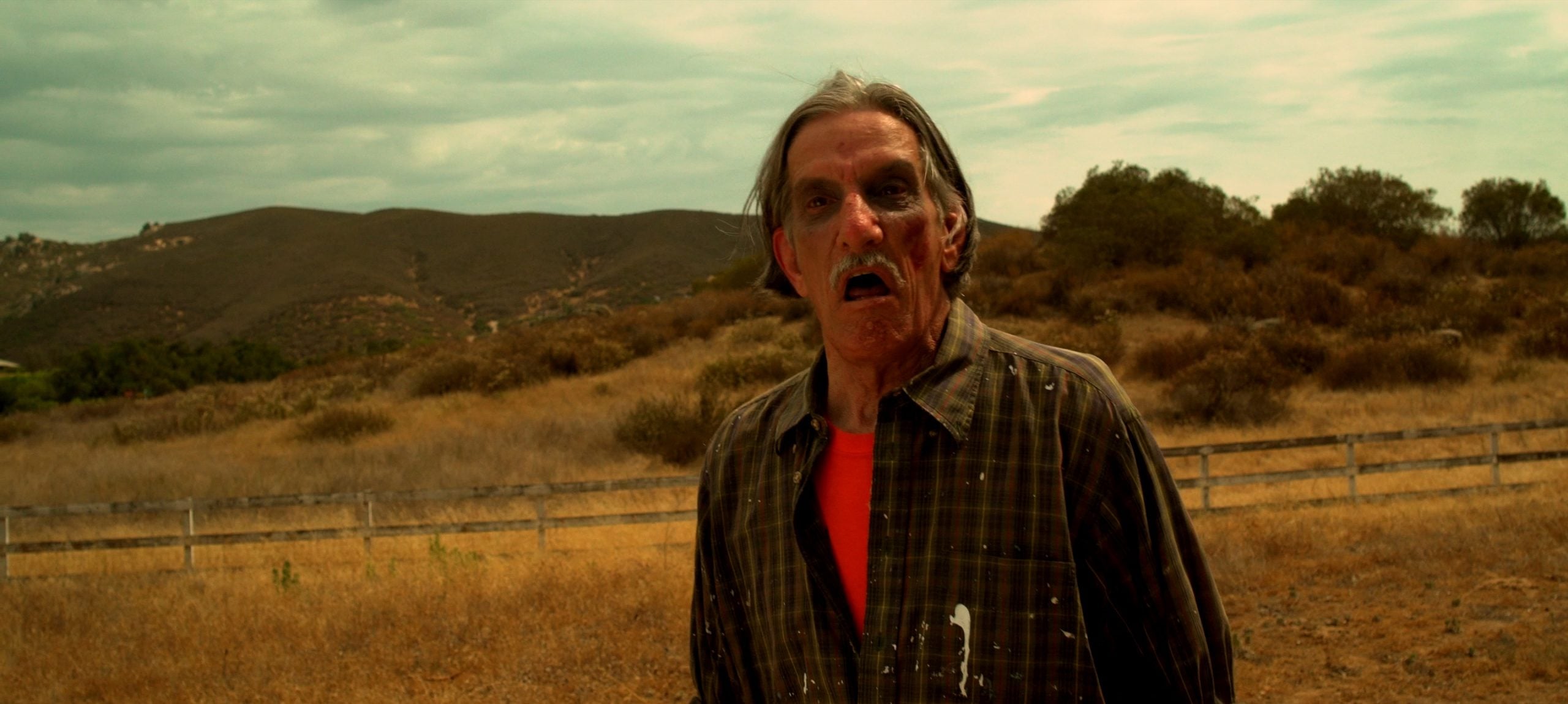 An older man with a surprised expression stands in a dry, grassy field with hills and a cloudy sky in the background.