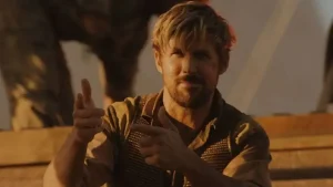 A man with blond hair and a beard sits outdoors, wearing a brown vest and shirt, making finger gun gestures with both hands.