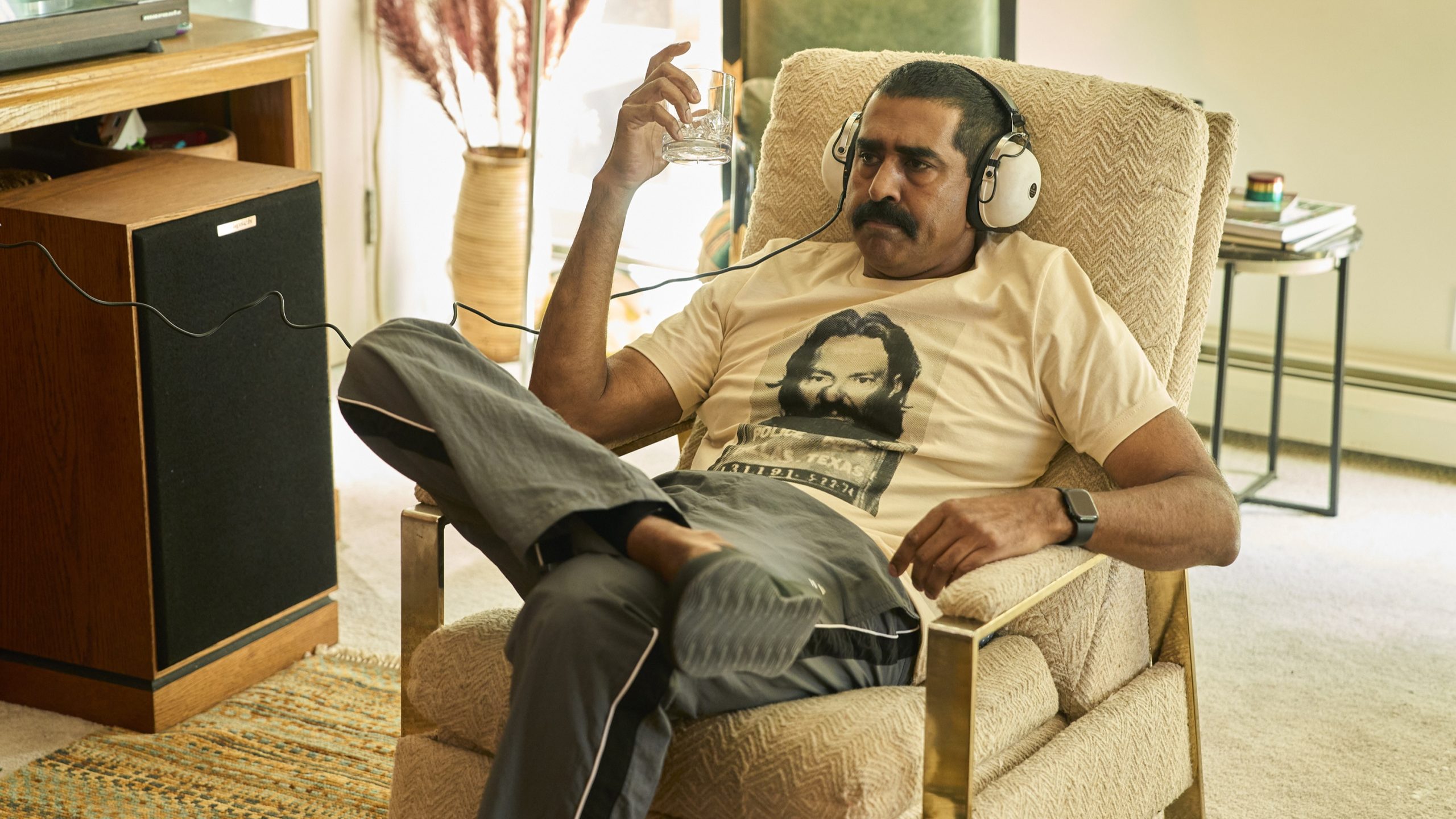 A man sits in an armchair wearing large headphones, holding a glass, and wearing a t-shirt with a bearded man’s face printed on it.
