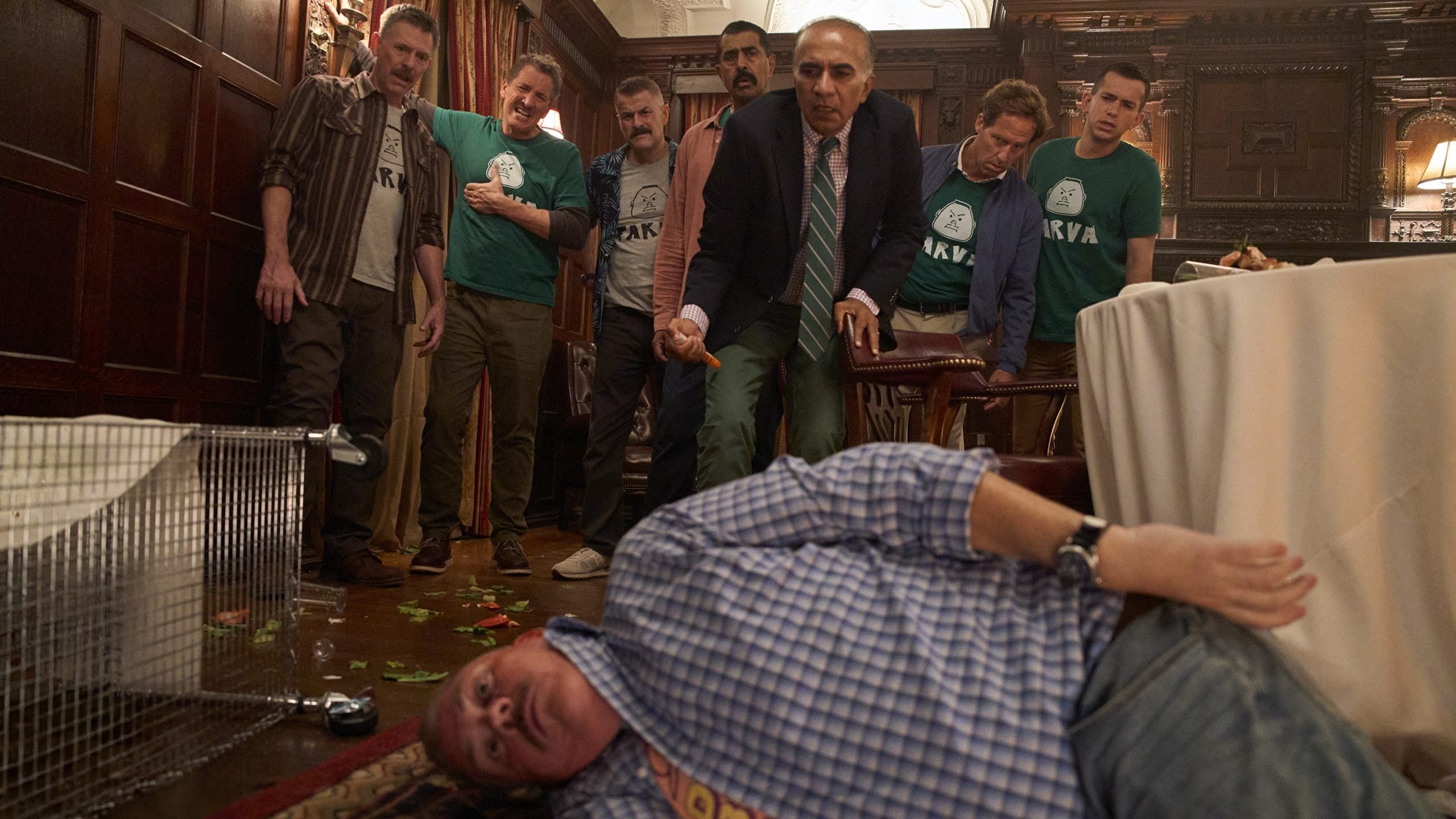 A group of men stand in a wood-paneled room, looking down at a man lying on the floor near an overturned table and scattered food.