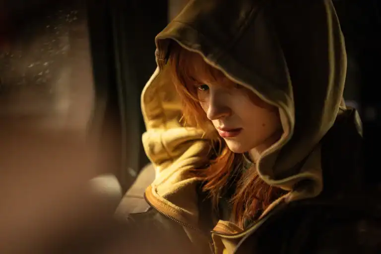 A person with red hair wearing a hooded jacket sits indoors, looking down with a serious expression. Warm light illuminates their face.