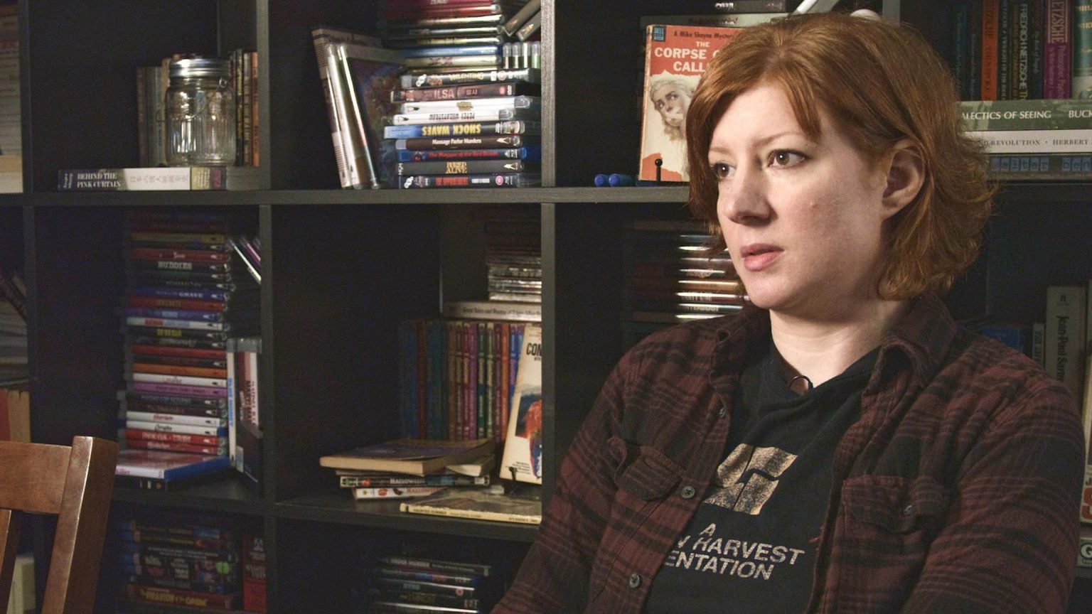 A person with short red hair sits indoors, facing right. Behind them is a dark bookshelf filled with books, DVDs, and a decorative jar.
