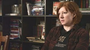 ‘Boutique: To Preserve And Collect’ Blu-Ray Review – Physical Media Gets Its Time In The Spotlight A person with short red hair sits indoors, facing right. Behind them is a dark bookshelf filled with books, DVDs, and a decorative jar.