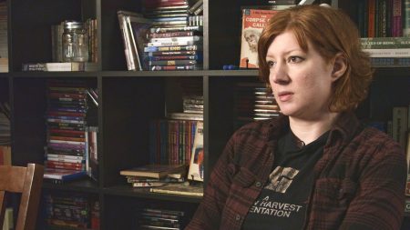 A person with short red hair sits indoors, facing right. Behind them is a dark bookshelf filled with books, DVDs, and a decorative jar.
