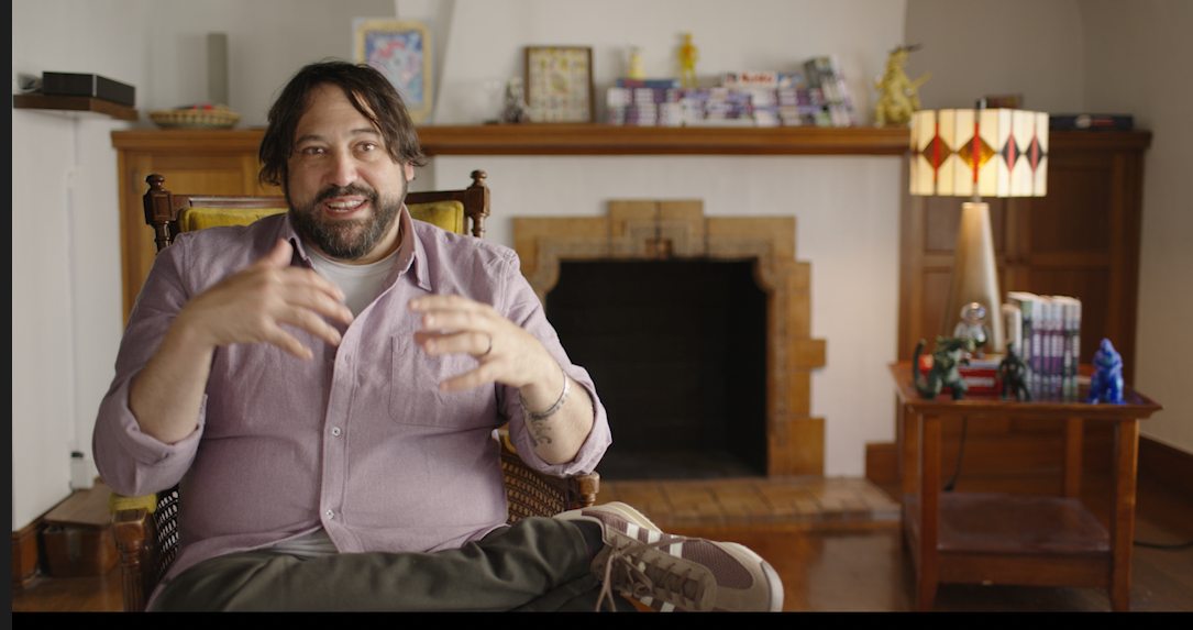 A man with long hair and a beard sits in an armchair, gesturing with his hands, in a living room with a fireplace and shelves filled with various items.