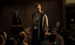 ‘The Samurai And The Prisoner’ Review – Finding A Way Out Of The Cycle Of Violence A man in traditional Japanese attire stands and speaks to a seated group in a dimly lit room; a suit of samurai armor is displayed in the background.