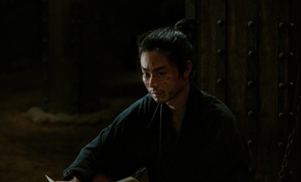 A man with a topknot and dark robe sits in a dim room, holding papers, with a serious expression on his face.