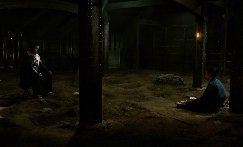 Two people sit on opposite sides of a dimly lit, earthen room with scattered rocks, wooden beams, and sparse furniture.