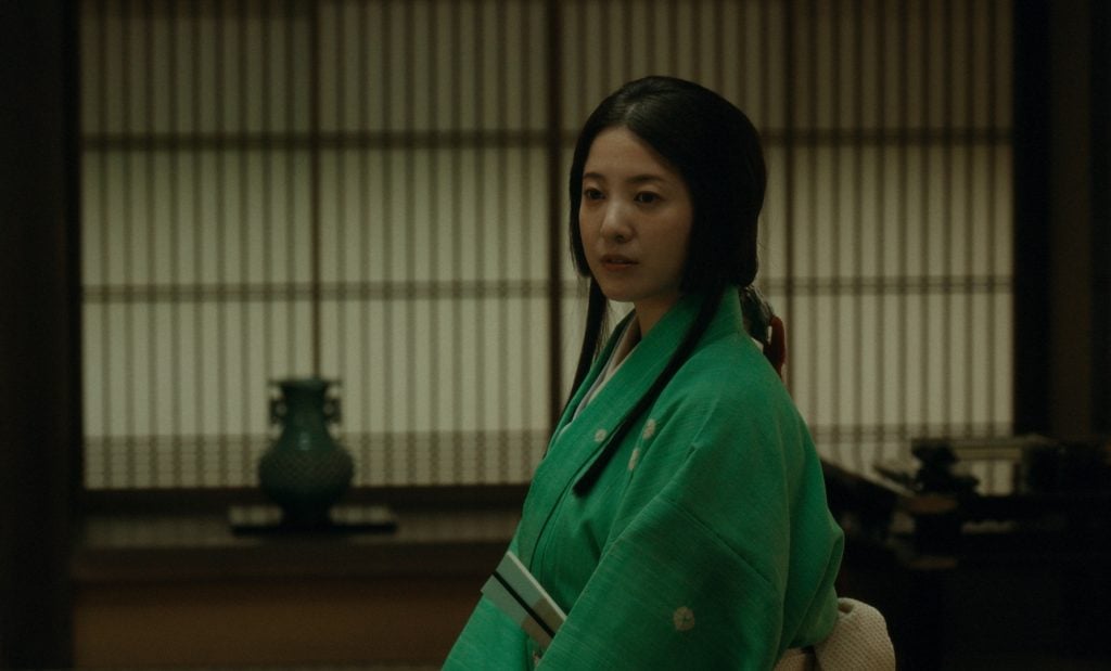 A woman in a green kimono sits indoors in front of traditional wooden latticed windows, with a green vase and other items visible in the background.
