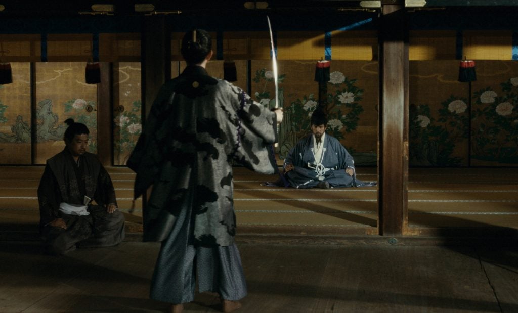 Three people in traditional Japanese clothing are in a wooden room; one holds a sword, one sits cross-legged facing him, and one observes from the side.