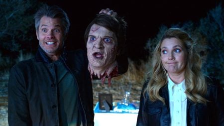 A man grins while holding a severed, realistic human head prop; a woman beside him looks unimpressed. They stand outside at night with a table in the background.