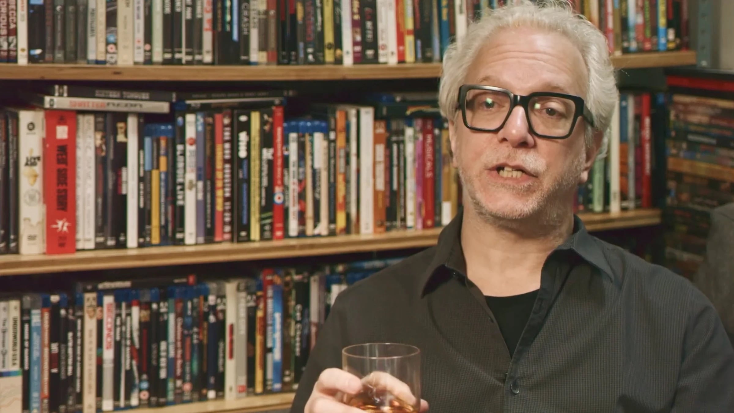 A man with gray hair and glasses holds a drink in front of shelves filled with DVDs or Blu-rays.