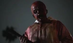 A person wearing a disturbing baby mask and blood-stained clothes holds a large object, possibly a weapon, against a dark, shadowy background.
