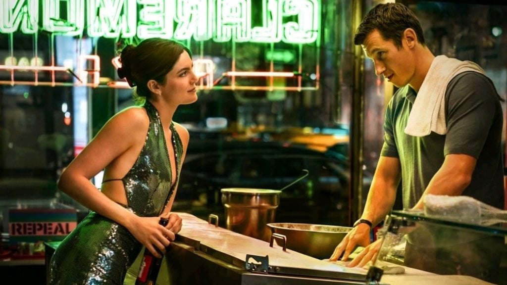 A woman in a shiny dress leans on a diner counter while talking to a man in a T-shirt with a towel over his shoulder. Neon signs glow in the background.