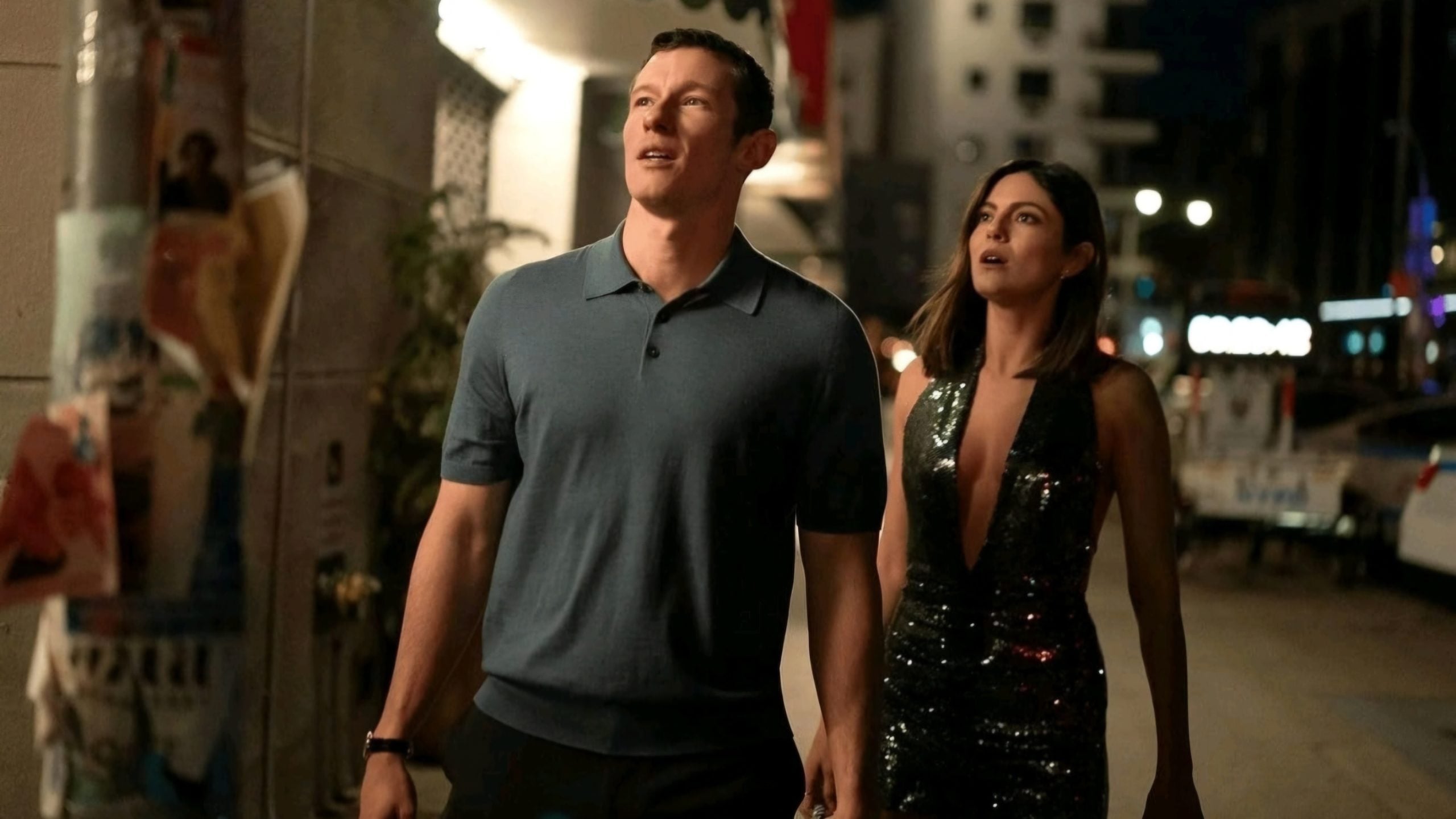 A man in a blue polo shirt and a woman in a sparkly, deep-neck dress walk outside at night, looking upward.