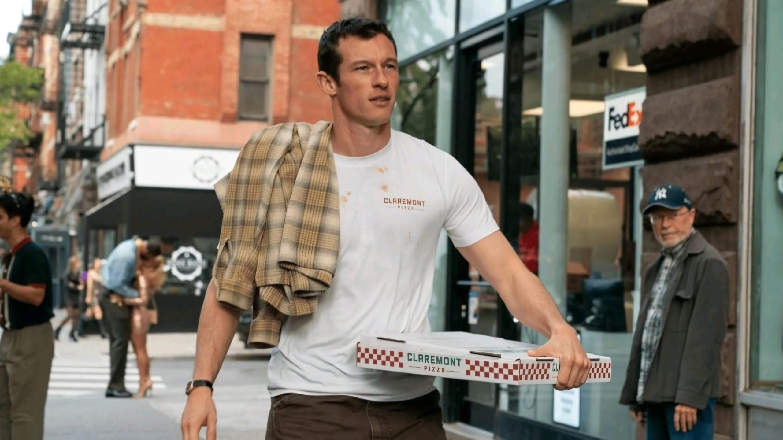 A man in a white shirt carries a pizza box and a plaid jacket over his shoulder while walking on a city sidewalk.