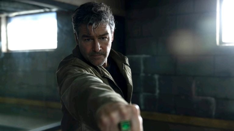 A man with gray hair and a stubble beard in a brown jacket points his fist forward, displaying a green ring, in a dimly lit concrete room.