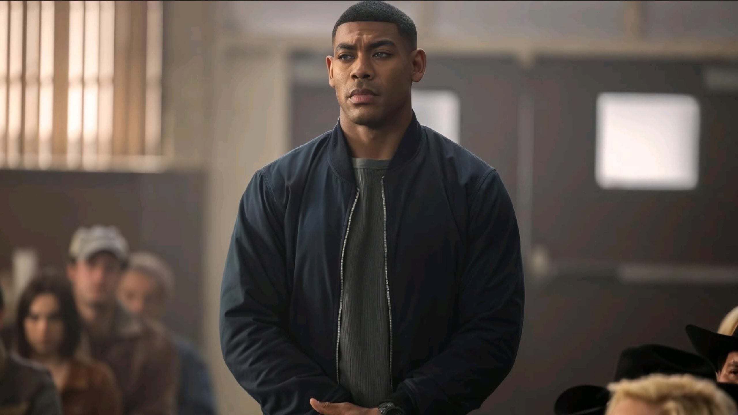 A man in a dark jacket stands indoors with hands clasped, looking serious, as several people sit in the background.