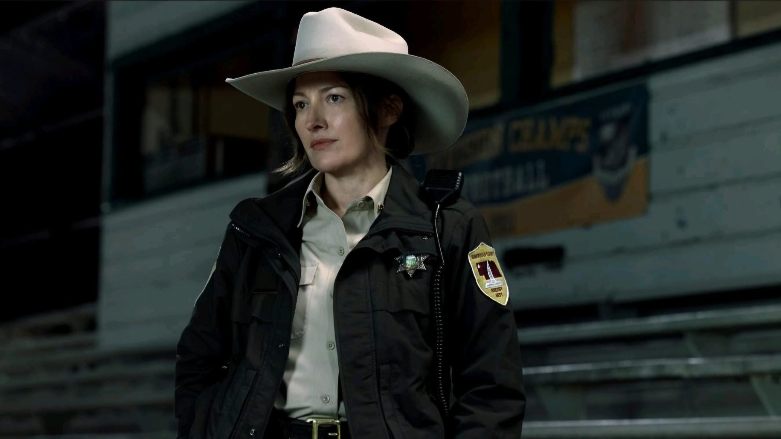 A sheriff in uniform and a white cowboy hat stands indoors with bleachers and a faded sign visible in the background.