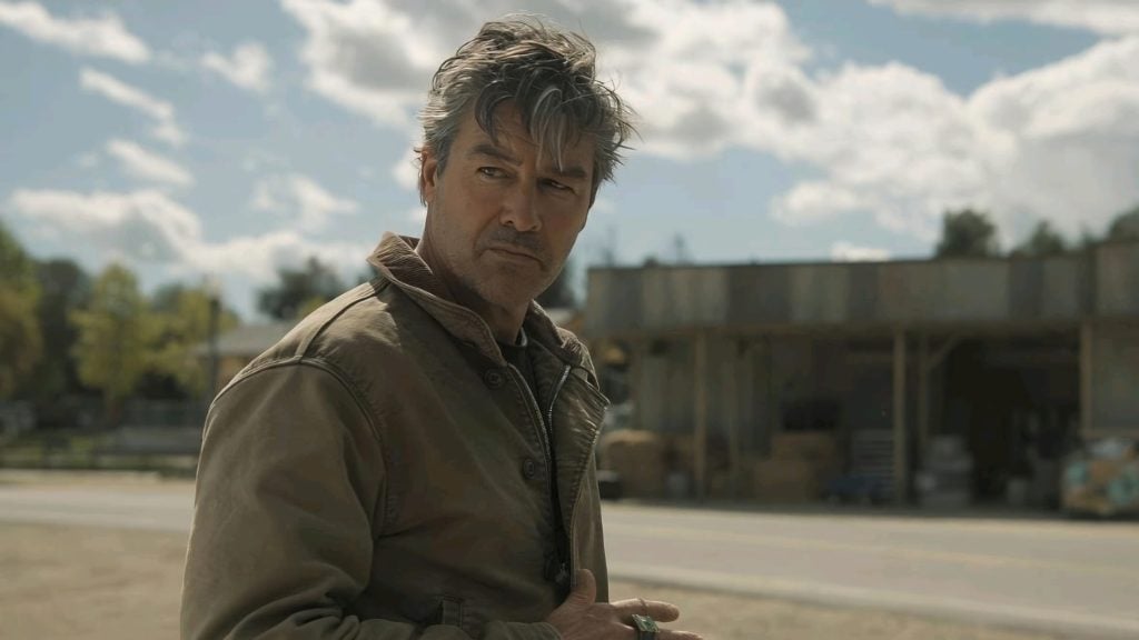 A man with gray hair in a brown jacket stands outside near a road, looking to the side. A weathered building and a cloudy sky are visible in the background.