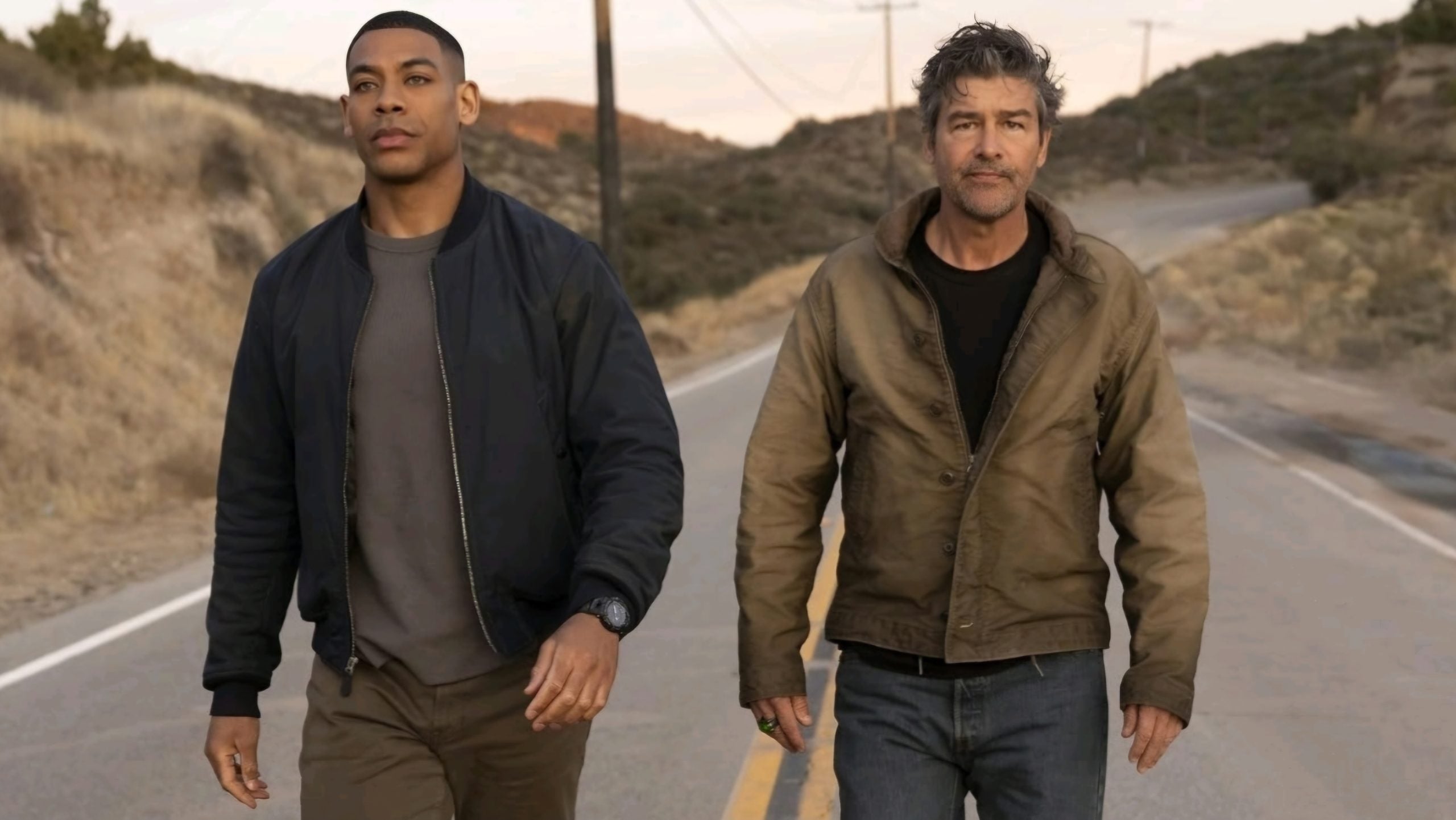 Two men walk down a rural road at sunset, both wearing jackets and looking ahead with serious expressions.
