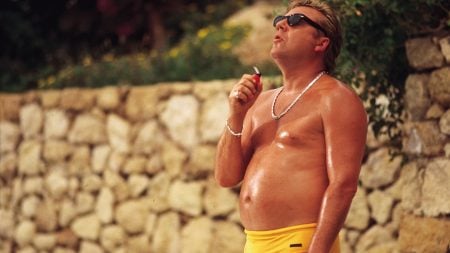 Second Sight Films Announces September Titles Including Works From Jonathan Glazer, Sam Waterston & More A man with blond hair, wearing sunglasses, a necklace, and yellow swim trunks, stands outdoors in front of a stone wall, holding a small object and looking up.