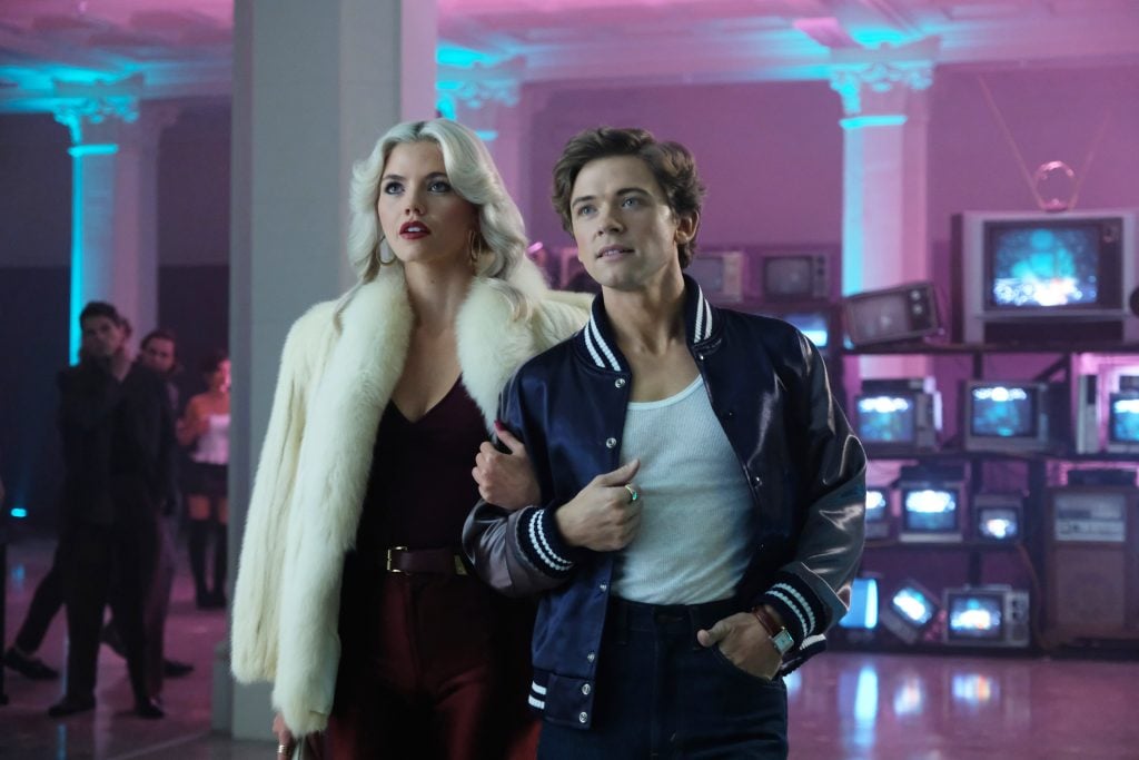 A woman in a white fur coat and a man in a varsity jacket walk arm-in-arm in a neon-lit room with stacked vintage TVs in the background.